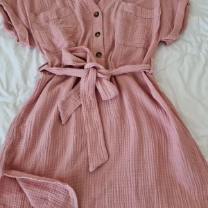 Skön rosa bomullsklänning - Oanvänd. Skön bomull. Med kort ärm, fickor, knytskärp och slitsar i sidorna.