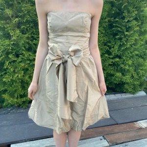 Festlig Klänning med rosett - Så fin beige/champagne färgad offshoulder klänning med ett tjockt band man knyter till en stor rosett på framsidan eller baksidan beroende på vad tycker bäst om 🎀 Har endast använt den en gång så den är som ny! Nyttpris var ca 1999kr och säljer 700kr