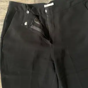 Svarta kostymbyxor från NLY TREND (Nelly.com) med utsvängda ben, försluts med en knapp och dragkedja. Två fickor framtill samt två fickor som detalj baktill.