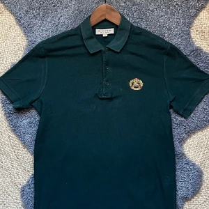 Burberry Piké Archive Logo - Burberry Piké i mossgrön färg, Plagget är köpt 2018 på NK, och är knappt använt. Plagget är del av archive collection, därav deras originella logga. Plagget är utav ett luftigt material, perfekt för golf och sommaraktiviteter. Köpt för 3500kr