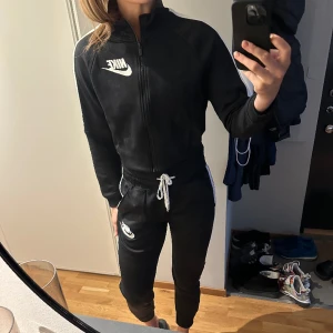 Nike-tracksuit - Set med tröja och byxor från Nike. Svart med vita ränder längs sidorna, Nike-logga både på tröjan och byxorna. Väldigt sparsamt använt, en handfull gånger kanske. 