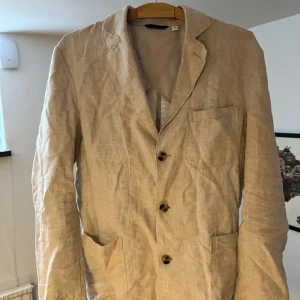 Beige kavaj - En beige kavaj med tre knappar framtill. Kavajen har en klassisk krage och en bröstficka på vänster sida. Den är tillverkad i ett lätt material, vilket gör den lämplig för vår- och sommarsäsonger. Linné köpt för 1200 självklart stryker jag sen innan 
