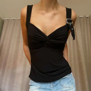 Topp med spänne - Snygg svart topp med ett spänne vid ena bandet. Jag har sytt upp den för hand i längden, kan behöva stärkas/sys om om man inte gillar avklippt slut. Annars bra skick, nästintill oanvänd. Står ej storlek men passar xs/s/m då den är stretchig