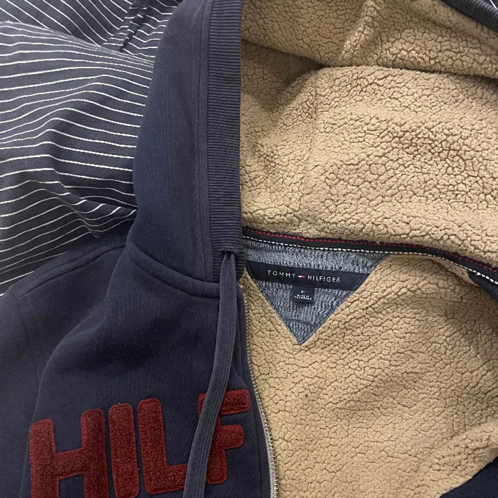 Marinblå huvtröja från Tommy Hilfiger med dragkedja, fickor och beige fluff inuti. Storlek M. Använd men i gott skick. Nött på muddarna, se bild tre. Inköpspris 850kr. Hoodies.