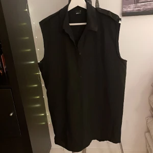 svart ärmlös skjorta - En svart ärmlös skjorta i lite tunnare material  Detalj med knapp på axlarna  Halvknäppning Storlek 40