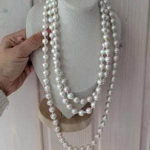 Ett elegant vitt pärlhalsband med stora, glänsande pärlor. Halsbandet är långt och kan bäras i flera lager för en fylligare look.