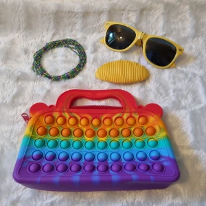 Flerfärgad handväska med pop-it design - En färgglad handväska med pop-it design i regnbågens färger. Väskan har ett handtag och en dragkedja för att stänga den. Säljer med hårclip, solglass ögon,och pärlor halsband. En perfekt present för barn.