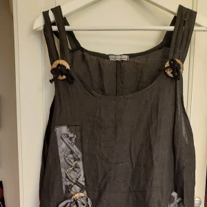 Linneklänning tunika storlek L - Snygg och unik klänning tunika med spännande detaljer med ringar i trä och band se foton. Den har en asymmetrisk form och är gjord i ett lätt och luftigt material. Perfekt för varma sommardagar.  Italiensk klänning i linne stl L