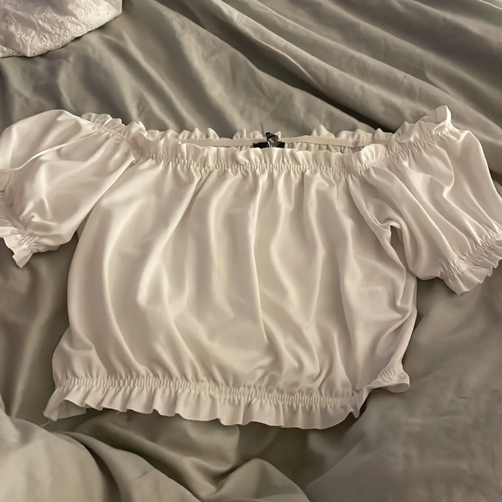 En vit off-shoulder topp med korta ärmar och elastiska band vid axlar, ärmslut och nederkant. Toppen har en lös passform och är tillverkad i ett lätt och bekvämt material.. Yläosat.