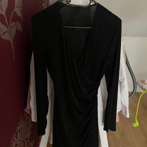Svart klänning  - Klänningen är i storlek M V-ringning med en omlottdetalj Lång ärm Rynkning i sidan Omlottkjol Minilängd Stretchigt, glansigt material! Jätte bra skick