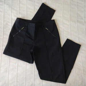 Svarta stretchiga kostymbyxor - ankellånga - storlek 38 - Svarta stretchiga byxor i en något kortare modell, från KappAhl. Dragkedjor i bensluten. 48% bomull, 48% nylon och 4% elastan. Storlek 38. Bara använda någon enstaka gång och i fint skick.