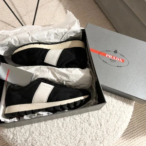 Prada sneakers - Prada sneakers som tyvärr bara ligger oanvända.  Serienr BE 6046 Svart tyg och mocka med vit revär och vit/svart sula.  Supersnygga både till dressad eller casual-looks. Orginalkartong medföljer.  Står strl 37,5 i men är 38,5-39 Pris kan diskuteras