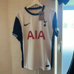 Tottenham 24/25 #18 LUCAS BERGVALL - En vit och blå Tottenham Hotspur fotbollströja från Nike. Tröjan har korta ärmar med blå detaljer på sidorna och ärmarna.. På baksidan finns namnet 'Bergvall' och nummer 18 tryckt i blått. Tröjan är en Reprint.
