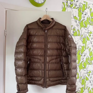 Brun dunjacka från Moncler - En brun dunjacka från Moncler med dragkedja framtill och två sidofickor med dragkedja. Modellen heter Acorus och är i storlek 4, något mellan en medium och large. Fint skick, äldre jacka. 