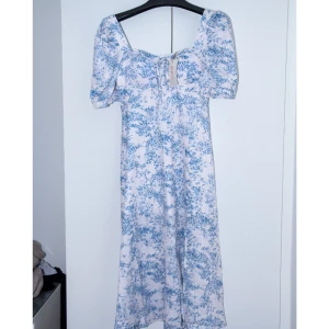 Blommig Midiklänning - En helt ny blommig sommarklänning som har korta puffärmar, hög midja, knytdetalj vid bysten och en slits längs ena sidan. Klänningen är gjort av polyester. Aldrig använd, lapparna är kvar.  Produktmått i cm  Byst: 86  Midja: 69  Höft: 99  Längd: 116 