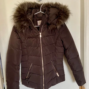 Vinterjacka - Brun vinterjacka med fejkpäls på luvan och gulddetaljer. Aldrig använd. Säljer för att den aldrig kommit till användning. 