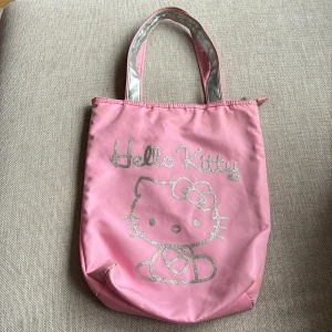 Väska Hello Kitty - Söt väska i form av en påse med motiv av Hello Kitty. Lite slitet på handtagen men annars i fint skick (kan skicka fler bilder) Stort fack med dragkedja och litet fack med dragkedja på insidan. Storlek ca 35x29 (utan handtag)