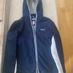 Jacka BOSS till pojke st 147-152 - Mörk blå jacka med luva, perfekt till våren av märket BOSS . Nyskick. Köpt på Nickis för ca 1300kr, knappt använd.  Finns i Bandhagen. Kan skickas, köparen står för frakten.