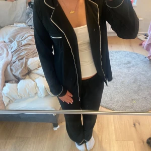 Svart pyjamasjacka med vita detaljer - Säljer ett svart pyjamas sett med vita detaljer. Den har en klassisk krage och knappar framtill. pyamasen är långärmad och har en avslappnad passform. Perfekt för mysiga hemmakvällar eller som en del av en pyjamasoutfit.