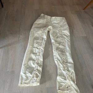 Beige byxor från & Other Stories - Säljer ett par snygga beige byxor från & Other Stories. De är i storlek 36 och har en cool snörningsdetalj längs benen. Byxorna är högmidjade och har en rak passform. Perfekta för både vardag och fest!