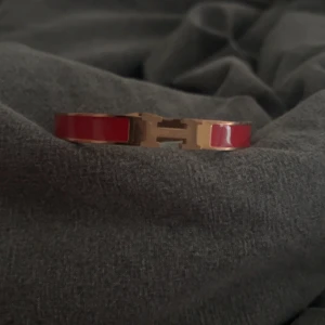 Rött armband med gulddetaljer - Säljer ett snyggt rött armband med gulddetaljer. Armbandet har en elegant design med en guldfärgad H-formad spänne. Perfekt för att lägga till en touch av färg och stil till din outfit. Passar bra till både vardag och fest!