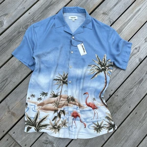 Kortärmad blå hawaiiskjorta - Säljer en härlig blå kortärmad hawaii-skjorta från Kappahl med ett tropiskt flamingomotiv. Skjortan är gjord av 100% viskos vilket gör den riktigt skön och luftig att ha på sig. Perfekt för sommarens alla fester eller bara en chill dag på stranden. Den har knappar framtill och en klassisk krage. Aldrig använd. Nypris 499 kr.