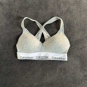 Grå sport-bh från Calvin Klein - Säljer en grå sport-bh från Calvin Klein. Den är i ett mjukt och stretchigt material med korsade axelband och en bred resår med Calvin Klein-loggan. Perfekt för träning eller vardagsbruk.