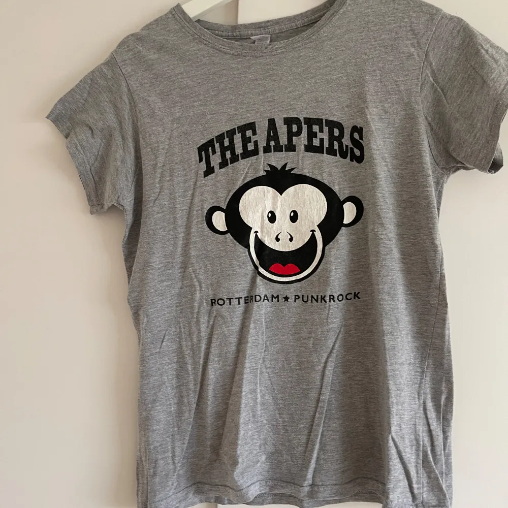 Säljer en grå t-shirt från Gildan i storlek XL. Den har ett coolt tryck med The Apers och en söt apa på framsidan, samt texten 'Rotterdam Punkrock'. På baksidan finns en liten logga från Asian Man Records. T-shirten är i bomull och är kortärmad. Perfekt för dig som gillar punkrock och vill ha en bekväm och stilren t-shirt!. T-paidat.