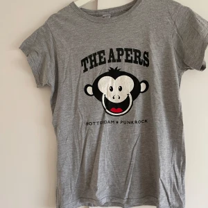 Grå t-shirt från Gildan med The Apers tryck - Säljer en grå t-shirt från Gildan i storlek XL. Den har ett coolt tryck med The Apers och en söt apa på framsidan, samt texten 'Rotterdam Punkrock'. På baksidan finns en liten logga från Asian Man Records. T-shirten är i bomull och är kortärmad. Perfekt för dig som gillar punkrock och vill ha en bekväm och stilren t-shirt!