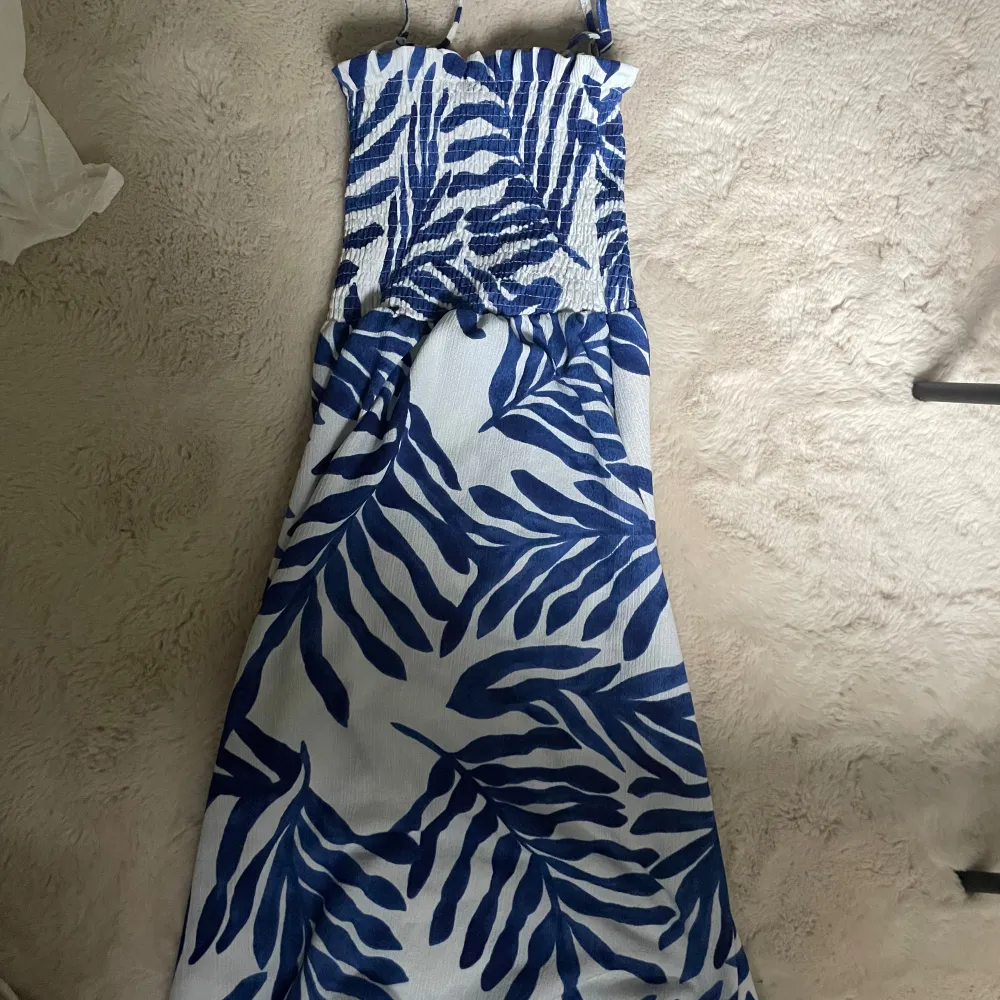 Säljer en superfin blåvit långklänning från H&M. Klänningen har ett snyggt bladmönster och är perfekt för sommaren. Funkar även som lång kjol och passar parfekt med en gullig topp. Den är ärmlös och har smala axelband. Materialet känns lätt och luftigt, vilket gör den idealisk för varma dagar. Perfekt för både vardag och fest! . Mekot.