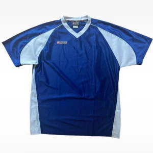 Blå fotbollströja från Lotto - Säljer en snygg blå fotbollströja från Lotto. Tröjan har en mörkblå bas med ljusblå paneler på sidorna och ärmarna. Den är kortärmad och har en V-ringad hals. Perfekt för fotbollsträning eller match!