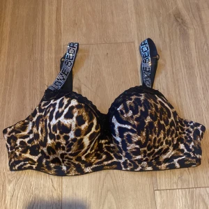 Leopardmönstrad bh med glittriga axelband - Säljer en snygg leopardmönstrad bh med glittriga axelband.. Den är knappt använd och har en fin spetsdetalj vid kuporna. Axelbanden är justerbara och bh:n stängs med hyskor i ryggen.