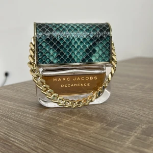 Marc Jacobs Decadence Parfym - Säljer en lyxig Marc Jacobs Decadence parfym. Flaskan är superfin med en grön ormskinnsdetalj på locket och en guldfärgad kedja som ger den en exklusiv känsla. Parfymen har en djup och sensuell doft som passar perfekt för speciella tillfällen. Flaskan är i glas och har en elegant design som ser ut som en liten handväska. Perfekt för dig som vill ha en doft som sticker ut!