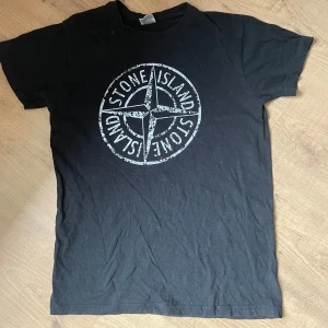 Svart t-shirt från Stone Island - Säljer en svart t-shirt från Stone Island med deras ikoniska logga tryckt på framsidan. T-shirten är i bomull och har korta ärmar. Perfekt för en casual look eller för att visa upp din stil på stan. Den är i välanvänt skick men fortfarande snygg och bekväm.