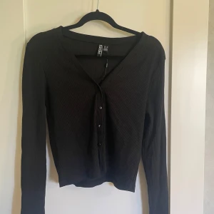 Svart ribbad kofta från Pieces - Säljer en snygg svart ribbad tröja som även går att använda som en kofta från Pieces. Den har en v-ringning och knappar framtill. Koftan är i nyskick och har långa ärmar.