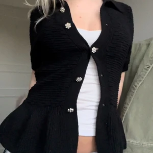 Svart kofta med pärlknappar - Säljer en snygg svart kofta med pärlknappar. Den har en figursydd passform och är perfekt för både vardag och fest. Koftan är kortärmad och har en fin struktur i tyget. Passar bra över en topp eller klänning.