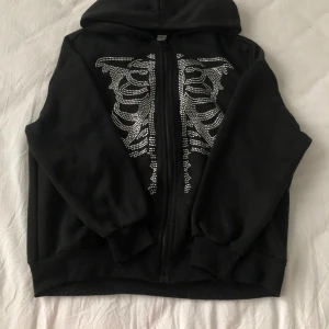 Svart hoodie med skelettmönster från Shein - Säljer en cool svart hoodie i storlek 13Y. Den har ett snyggt skelettmönster i silverfärgade nitar på framsidan och en dragkedja. Perfekt för höst och vinter! 🖤