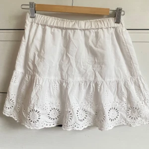 Vit kjol med broderade detaljer - Säljer en superfin vit kjol med elastisk midja och vackra broderade detaljer längst ner. Kjolen är kort och perfekt för sommaren. Den har en volangkant som ger en söt och feminin touch. Passar perfekt till både vardag och fest! Står ej storlek men passar XS  från kappahl tror det kan vara barn storlek 158?🥰
