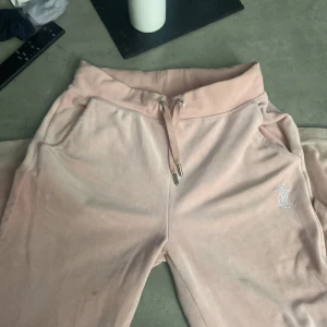 Rosa mjukisbyxor från Juicy Couture - Säljer ett par supermysiga rosa mjukisbyxor från Juicy Couture. byxorna  är dekorerade med glittriga stenar som bildar texten 'Juicy' på baksidan och en logga på framsidan. Perfekta för en chill dag hemma eller en avslappnad dag ute. 🩷