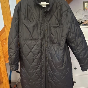 Svart quiltad jacka - En svart quiltad jacka med långa ärmar och knappar framtill. Jackan har två stora bröstfickor och en rak passform. Har aldrig används! 