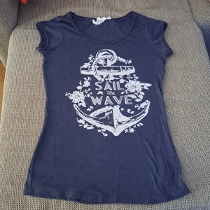 Mörkblå t-shirt med tryck - En mörkblå t-shirt med ett vitt tryck av ett ankare och texten 'Sail the Wave'. T-shirten har korta ärmar och en rund halsringning. oanvänd. 100% ny