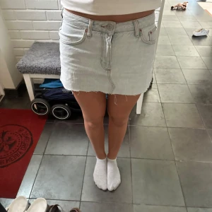 Ljusblå jeanskjol från BDG Urban Outfitters - En ljusblå jeanskjol från BDG Urban Outfitters. Kjolen har en kort längd och är tillverkad i denim med en rå kant nedtill. Den har klassiska fem fickor och stängs med knapp och dragkedja framtill.