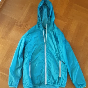 Blå vindjacka från C&A - En blå vindjacka från C&A med huva och dragkedja. Jackan har två framfickor med dragkedjor och elastiska muddar vid ärmslut och nederkant.