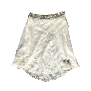 Vit kjol med paljetter  - Vit kjol med asymmetrisk design och paljetter längs kanterna. Kjolen har en broderad midja med pärlor  i guld och silver samt en knytning med pärl-tofsar