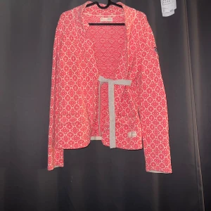 Rosa mönstrad jacka från Odd Molly - En rosa mönstrad jacka från Odd Molly med ett vitt geometriskt mönster. Jackan har långa ärmar och ett broderat märke på vänster ärm med texten 'Rise Above'. Den har en knytning i midjan och är tillverkad i ett stickat material. liten fläck på ena ärmen 