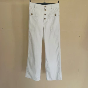 Vita byxor med knappar - Ett par vita byxor med hög midja och raka ben. Byxorna har fem knappar framtill och två dekorativa fickor med knappar på framsidan. De är tillverkade i ett randigt material.