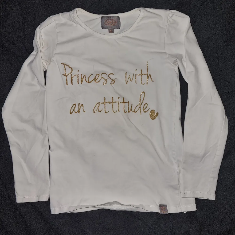 En vit långärmad tröja med texten 'Princess with an attitude' i guldglittrig skrift. Tröjan har en rund halsringning och är tillverkad i ett mjukt material. Från Creamie i stl 146. Neuletakit & Villapaidat.