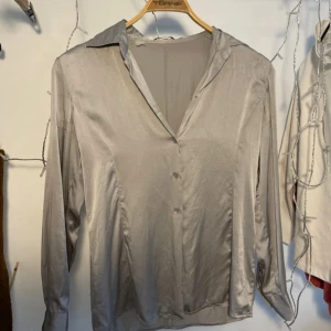 Skjorta / blus i 100 % silke - Snygg silvrig skjorta i 100 % silke. Sömmen är lite trasig bak på ena ärmen samt lite sliten i söm bak, se bilder. Översta knappen saknas men det finns en extraknapp som lätt kan sys på! Gansk liten i storleken.