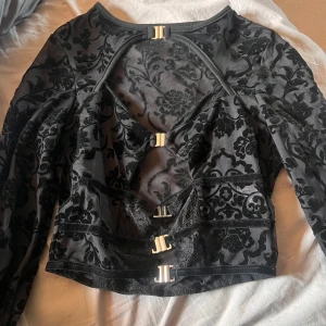 Svart blus med spetsdetaljer och spännen - En elegant svart blus med spetsdetaljer och 4 dekorativa spännen framtill. Blusen har ett blommigt mönster och långa ärmar. Den är perfekt för festliga tillfällen. aldrig använd 
