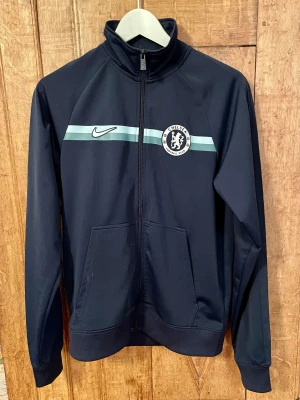 NIKE Chelsea track top i superfint skick - Säsongen är i gång och de blåa behöver ditt stöd! Finns inget bättre sätt än att flexa denna tracktop! Snygg stålblå mörkblå med Chelsea-loggan och Nike swooshen över bröstet. Två fickor fram. Fint skick, inga skavanker, knappt använd. Passar S el M. 